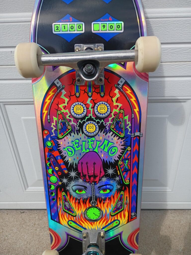 Santa Cruz Delfino skateboard review - A Delfino trick skateboard deck with vibrant pinball-inspired graphics, leaning against a white garage door.